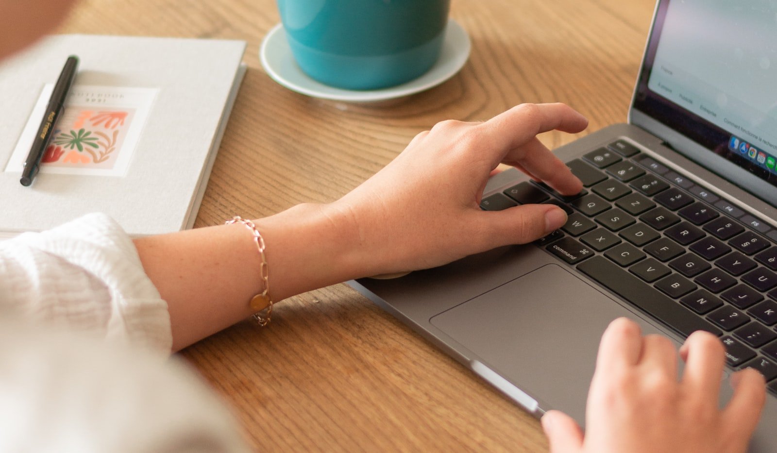 A hand typing on a laptop keyboard at a wooden desk beside a notebook and a cup of coffee