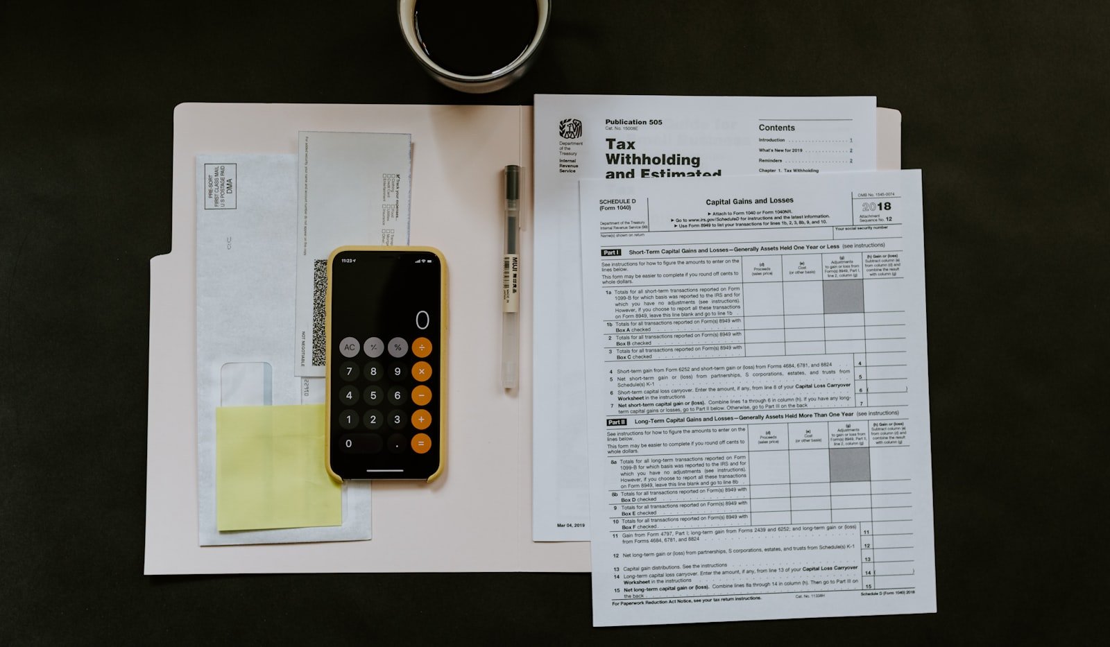 A phone calculator sitting on a folder of tax and financial paperwork beside a pen and a cup of coffee, viewed from above as someone works through a bill