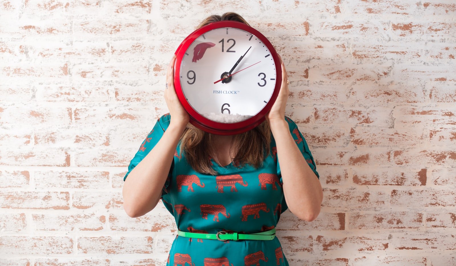 A woman holding a large red rimmed wall clock in front of her face while standing against a painted brick wall, its hands already well past the hour