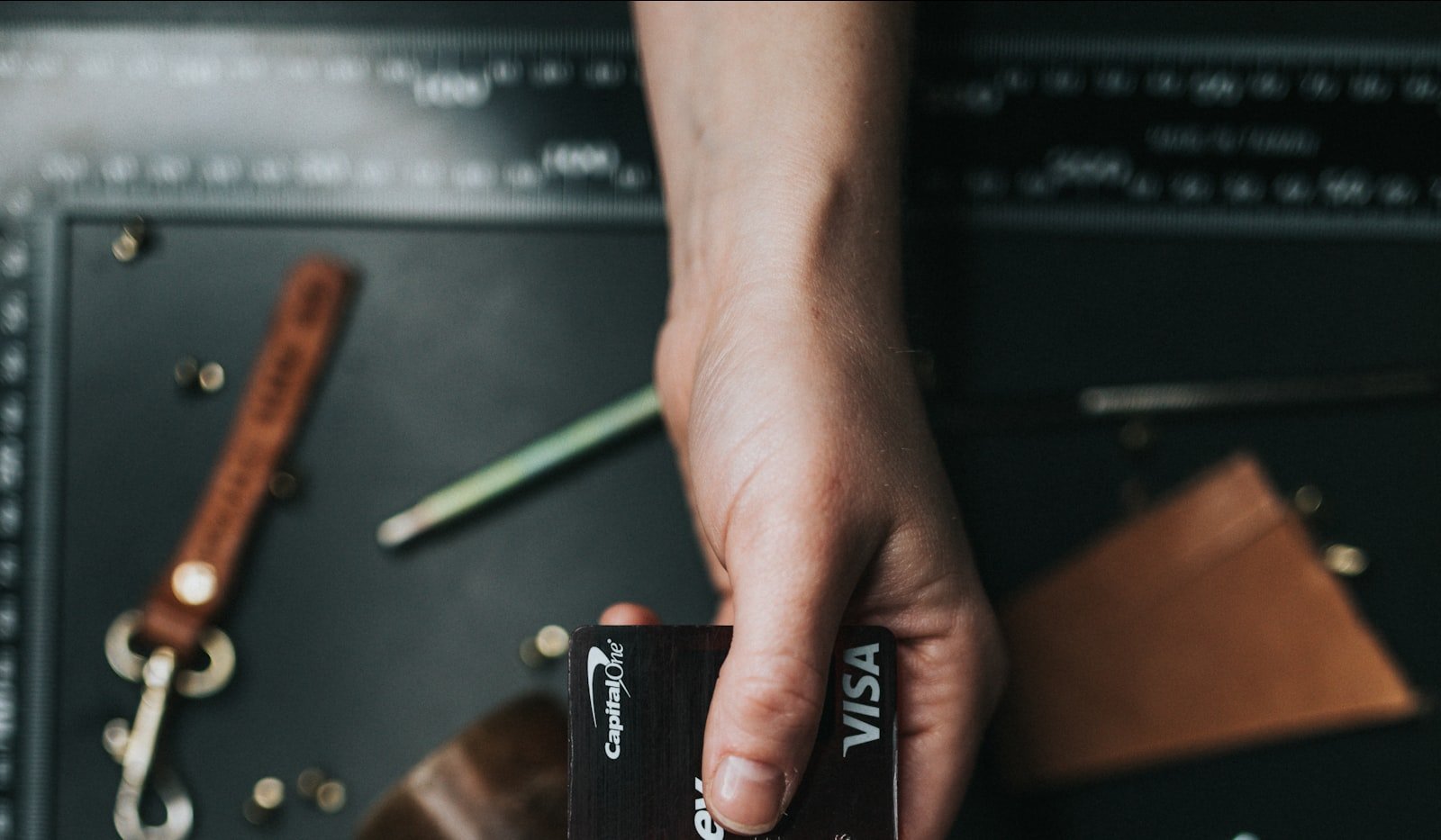 A hand holding out a Capital One Visa card over a dark leather desk with a pencil and keys nearby